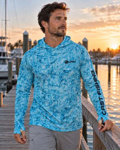 Man wearing a CastThreads Aqua Camo BreezeTec hooded performance shirt with CAST FISHING branding on the left sleeve at sunrise in a marina.